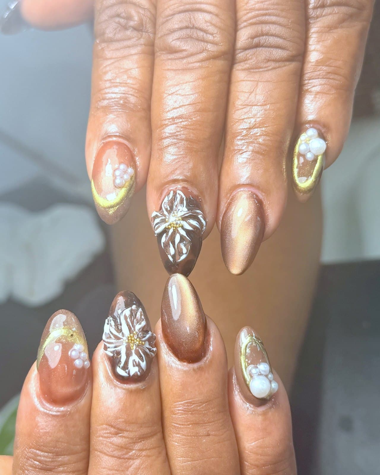 Pose en gel brun cat-eye avec fleurs 3D, chrome doré et perles, réalisée chez Couca & Co. Beauty